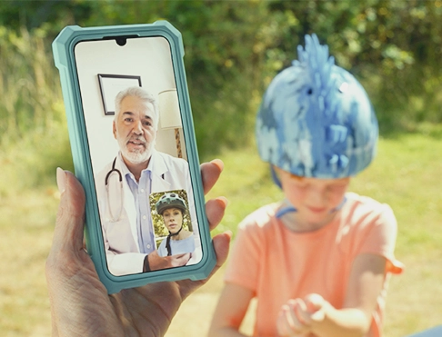 A cell phone video call with a doctor and a kid wearing a helmet looking over her arm in the background